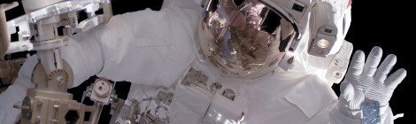 Ein Mensch in einem dicken, weißen Raumanzug winkt bei einem Außenbordeinsatz im Weltall in die Kamera. Das Gesicht ist nicht zu sehen, weil das Visier des Helms verspiegelt ist.