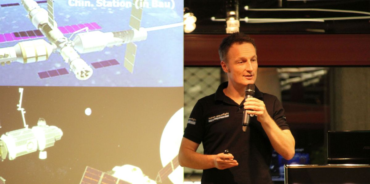ESA-Astronaut Matthias Maurer hält einen Vortrag. Im Hintergrund sieht man auf seinen Vortragsfolien Raumstationen im Erdorbit und im Mondorbit.