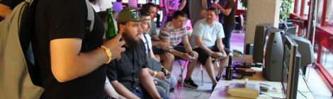 Drei Spielestationen des Retrogaming-Abends. Rechts stehen ein Flachbildschirm und zwei Röhrenmonitore auf zusammengestellten Tischen. Vor den Monitoren befinden sich drei Retro-Spielekonsolen und mehrere Spielekartuschen. Links sitzen sieben junge Erwachsene auf Stühlen nebeneinander, bedienen Gaming-Controller und blicken auf die drei Bildschirme. Drumherum und im Hintergrund sieht man weitere Erwachsene sich unterhalten, zugucken oder spielen.