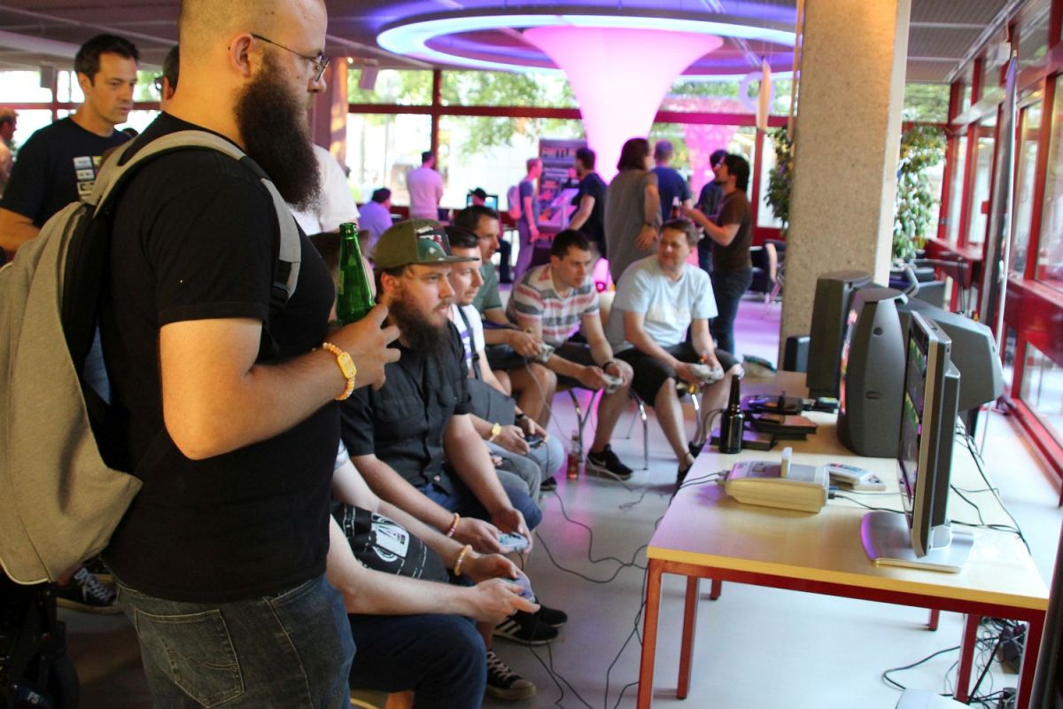 Drei Spielestationen des Retrogaming-Abends. Rechts stehen ein Flachbildschirm und zwei Röhrenmonitore auf zusammengestellten Tischen. Vor den Monitoren befinden sich drei Retro-Spielekonsolen und mehrere Spielekartuschen. Links sitzen sieben junge Erwachsene auf Stühlen nebeneinander, bedienen Gaming-Controller und blicken auf die drei Bildschirme. Drumherum und im Hintergrund sieht man weitere Erwachsene sich unterhalten, zugucken oder spielen.