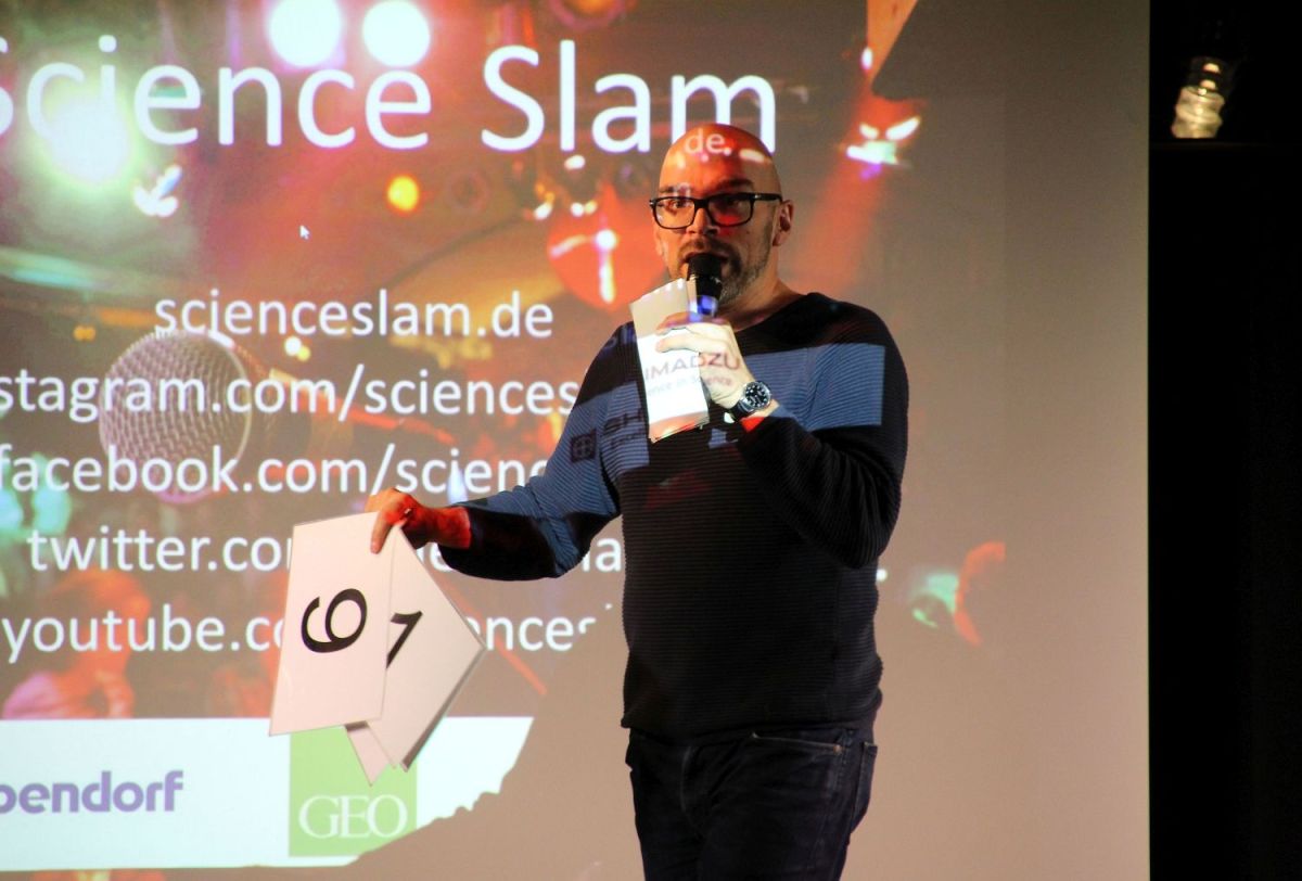 Der Moderator vor der Leinwand, die die Science-Slam-Startfolie zeigt. Der Moderator spricht in sein Mikrofon und hält ein Set Wertungskarten in der rechten Hand.