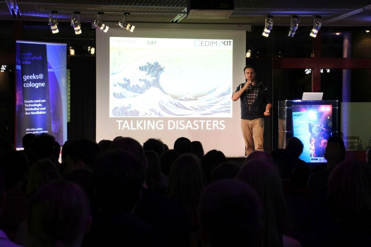 Blick auf die Bühne mit der Leinwand. Auf der Leinwand sind hohe Meereswellen zu sehen und die Worte "TALKING DISASTERS". Ein junger Mann steht auf der Bühne und spricht in ein Mikrofon.