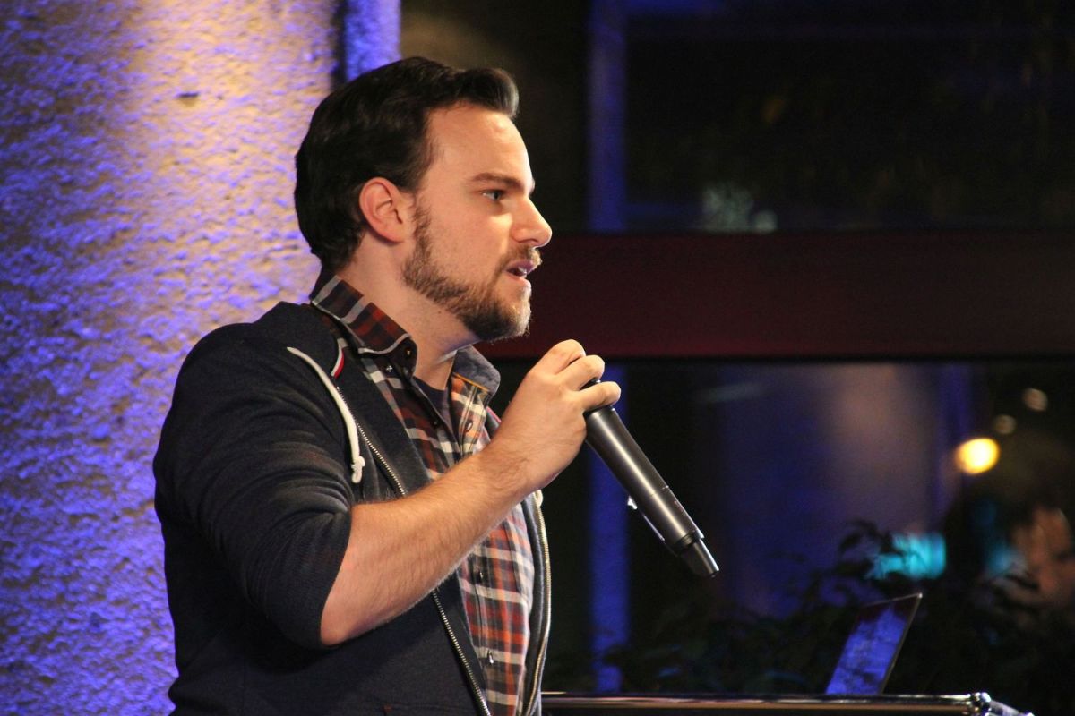 Ein junger Mann von der Seite fotografiert. Er spricht in ein Mikrofon.