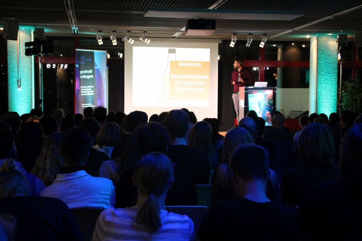 Blick auf die Bühne mit der Leinwand. Auf der Leinwand ist eine Bierflasche zu sehen und eine Warnmeldung. Neben der Leinwand steht junger Mann und hält eine Bierflasche in der Hand.