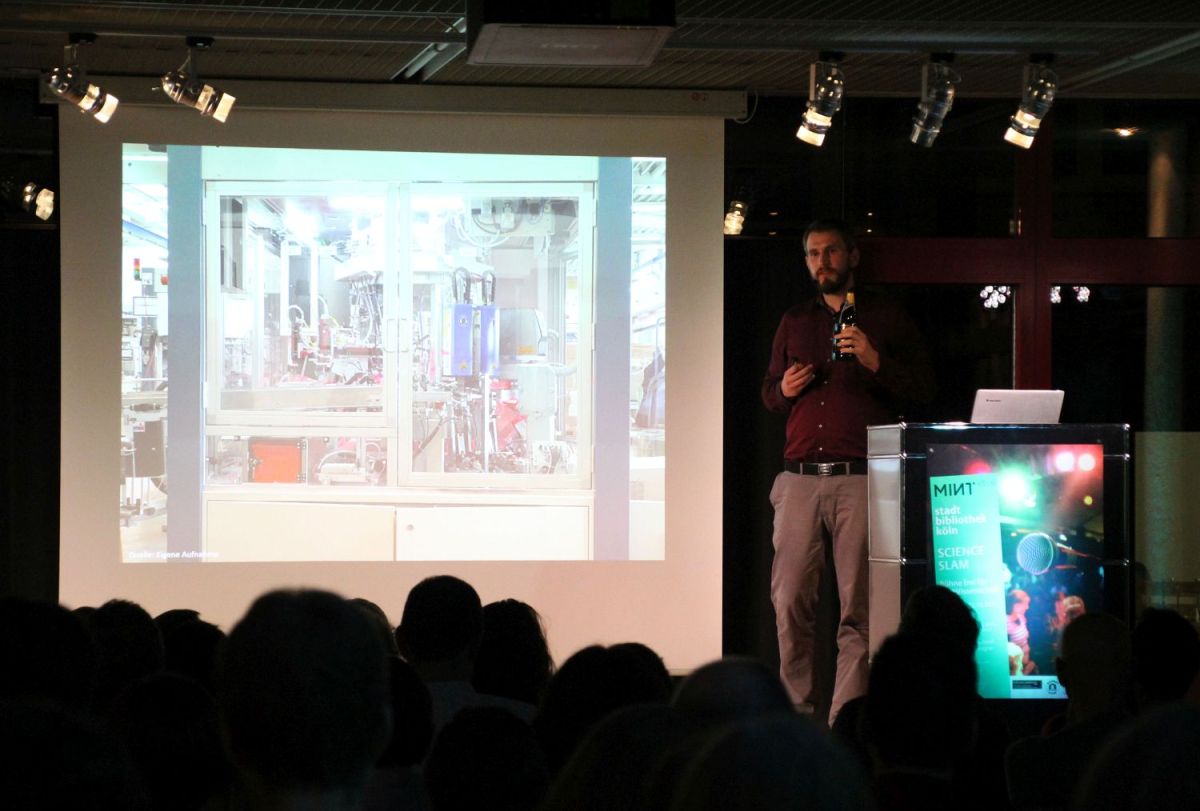 Blick auf die Bühne mit der Leinwand. Auf der Bühne steht ein junger Mann. Er hält eine Bierflasche hoch und wendet sich an das Publikum.