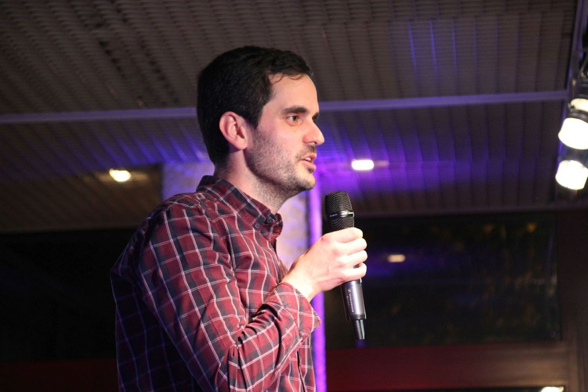 Ein junger Mann von der Seite fotografiert. Er spricht in ein Mikrofon.