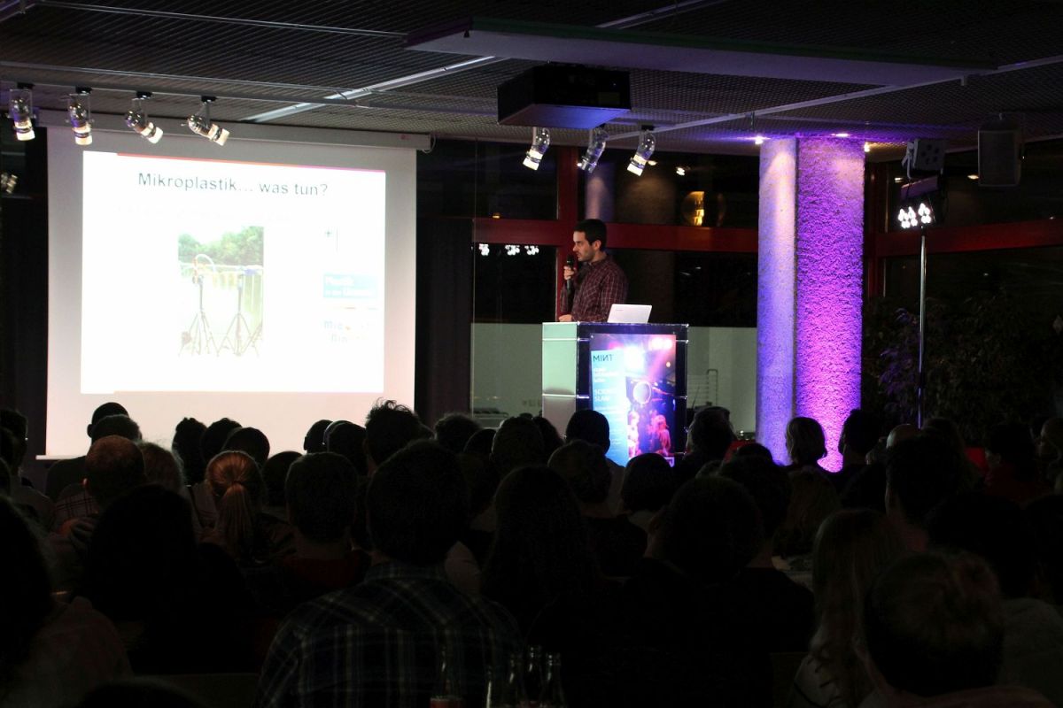 Blick auf die Bühne. Ein junger Mann spricht in ein Mikrofon und schaut zur Leinwand. Im Vordergrund sitzt das Publikum im Dunkeln.