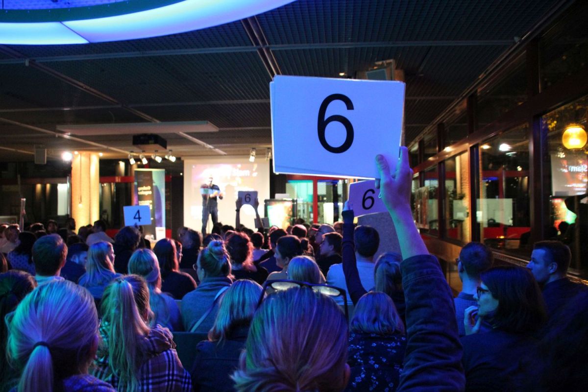 Eine der Wertungsphasen des Science Slams. Im Vordergrund die blau beleuchteten Hinterköpfe des Publikums. Vier Personen aus dem sichtbaren Publikum halten Wertungskarten hoch. Auf der Bühne notiert der Moderator die Wertungen.