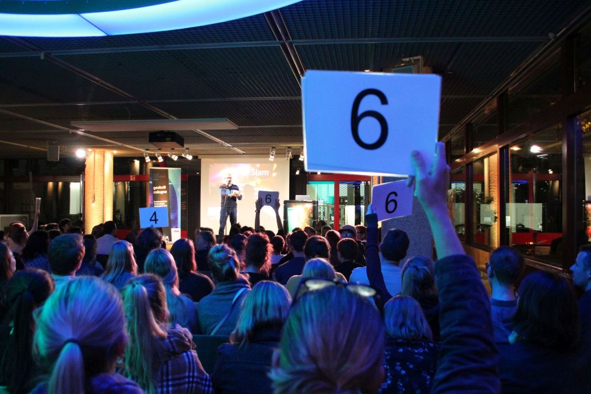 Eine der Wertungsphasen des Science Slams. Im Vordergrund die blau beleuchteten Hinterköpfe des Publikums. Vier Personen aus dem sichtbaren Publikum halten Wertungskarten hoch. Auf der Bühne notiert der Moderator die Wertungen.