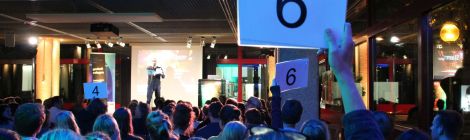 Eine der Wertungsphasen des Science Slams. Im Vordergrund die blau beleuchteten Hinterköpfe des Publikums. Drei Personen aus dem sichtbaren Publikum halten Wertungskarten hoch. Auf der Bühne notiert der Moderator die Wertungen.