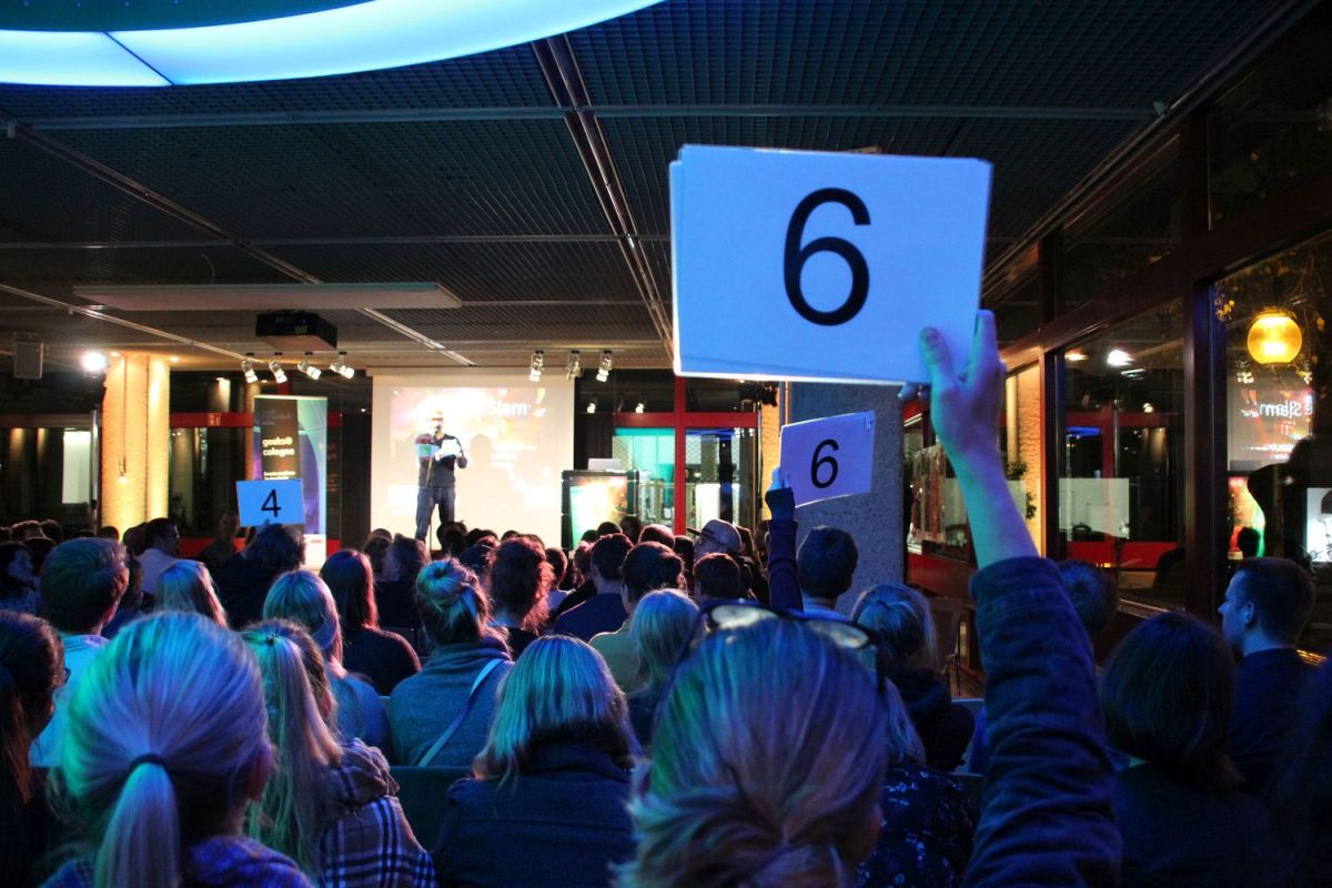 Eine der Wertungsphasen des Science Slams. Im Vordergrund die blau beleuchteten Hinterköpfe des Publikums. Drei Personen aus dem sichtbaren Publikum halten Wertungskarten hoch. Auf der Bühne notiert der Moderator die Wertungen.