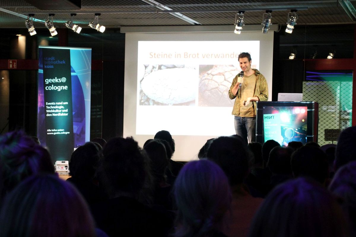 Blick auf die Bühne. Ein junger Mann spricht in ein Mikrofon. Hinter ihm befindet sich die Leinwand mit seiner Präsentation.