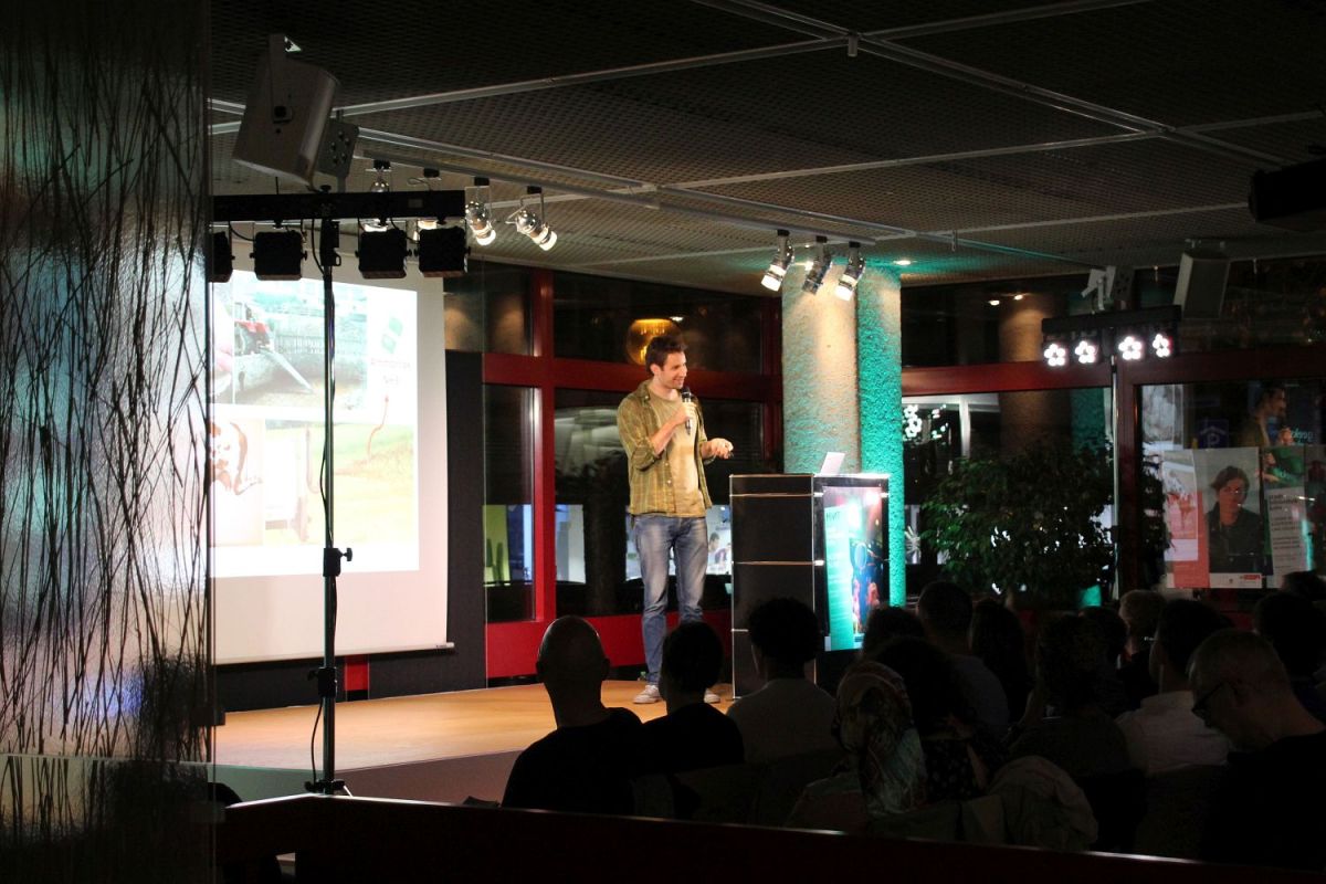 Links befindet sich die beleuchtete Bühne mit der Leinwand. Ein junger Mann steht auf der Bühne und spricht in ein Mikrofon. Rechts in der Dunkelheit sieht man das Publikum sitzen.