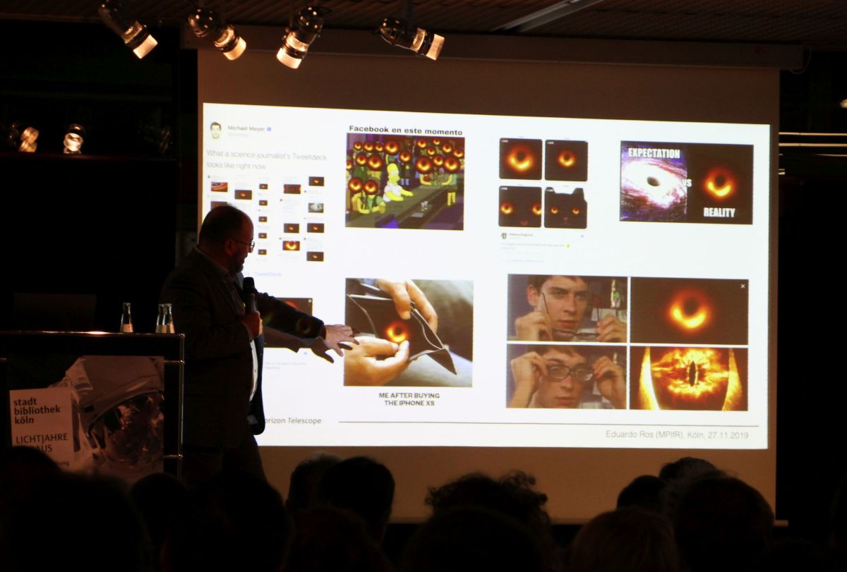 Veranstaltungsbühne der Zentralbibliothek. Leinwand mit Vortragsfolie. Darauf mehrere Memes mit dem Bild des Schwarzen Lochs. Zum Beispiel ein Schwarzes Loch in der Brieftasche. Oder eine Gegenüberstellung des von der Öffentlichkeit erwarteten spektakulär detaillierten Fotos eines Schwarzen Lochs mit dem tatsächlichen, sehr verschwommenen Ring. Links daneben der Vortragende.