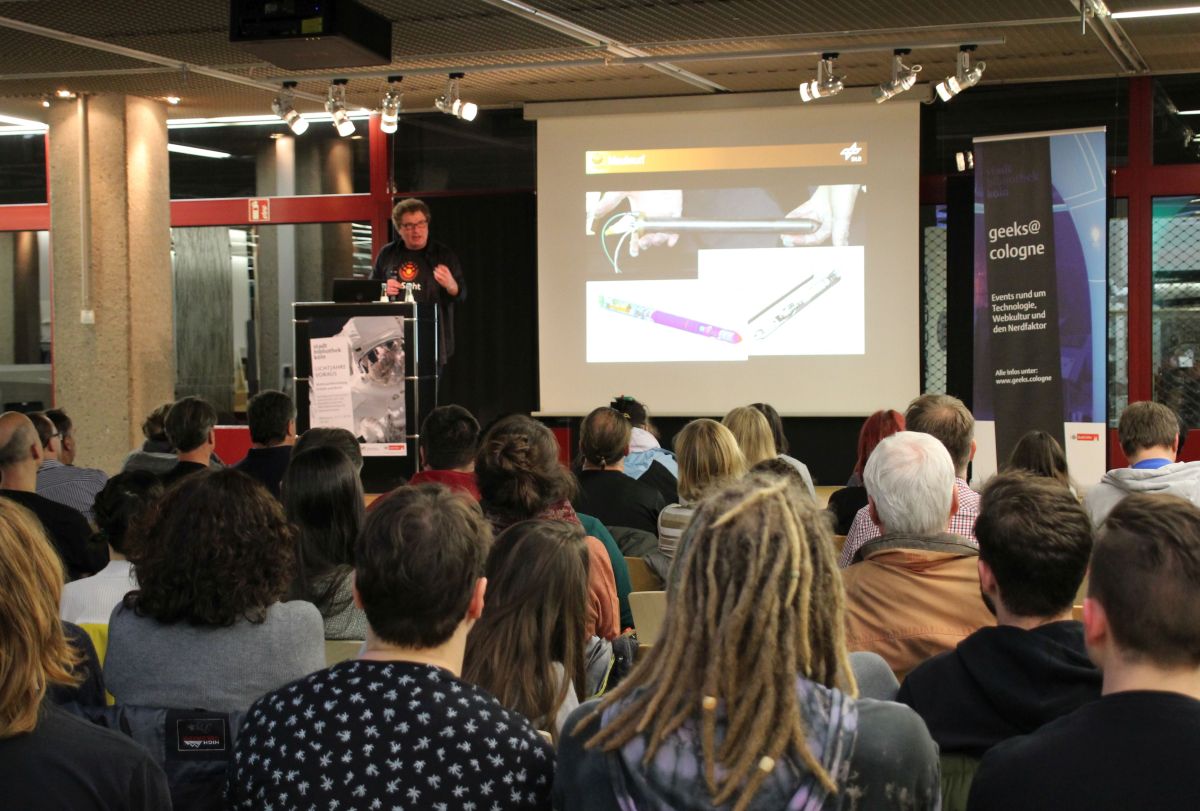 Veranstaltungsbühne der Zentralbibliothek. Leinwand mit Vortragsfolie. Links daneben der Vortragende. Publikum auf Stühlen im Vordergrund.