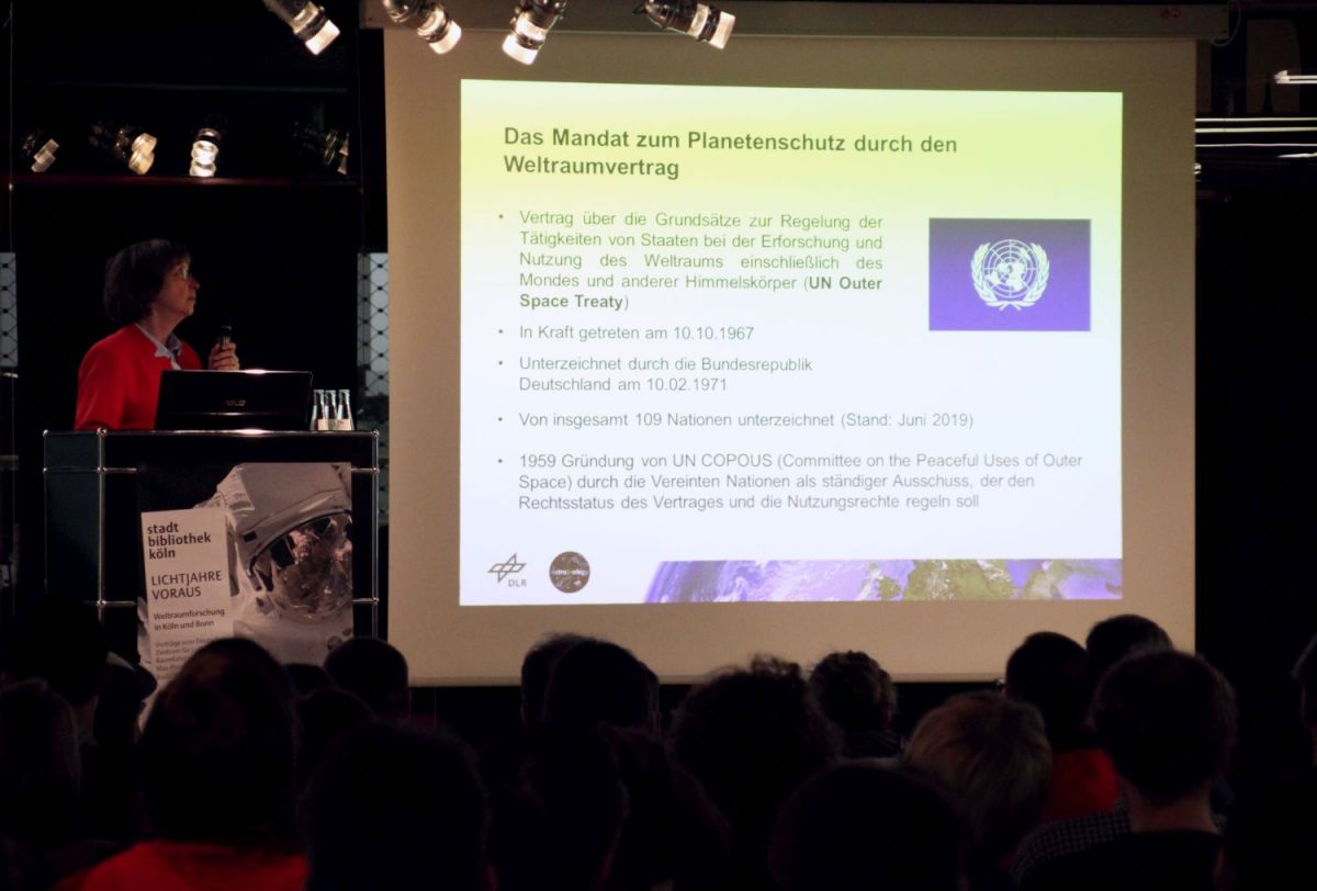 Veranstaltungsbühne der Zentralbibliothek. Leinwand mit Vortragsfolie. Links daneben die Vortragende. Publikum auf Stühlen im Vordergrund.