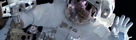 Ein Mensch in einem dicken, weißen Raumanzug winkt bei einem Außenbordeinsatz im Weltall in die Kamera. Das Gesicht ist nicht zu sehen, weil das Visier des Helms verspiegelt ist.