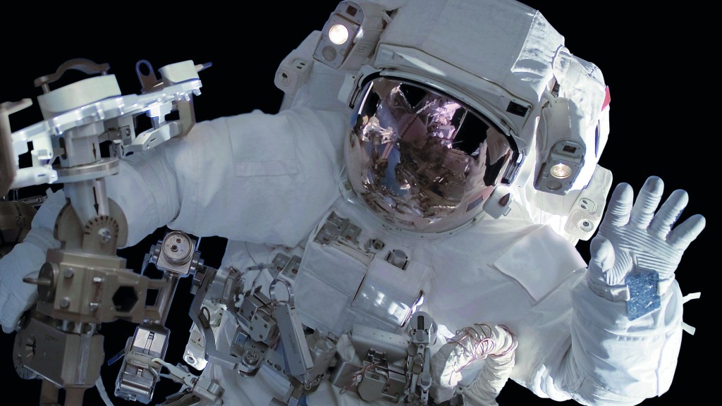 Ein Mensch im Raumanzug winkt bei einem Außenbordeinsatz im Weltall in die Kamera