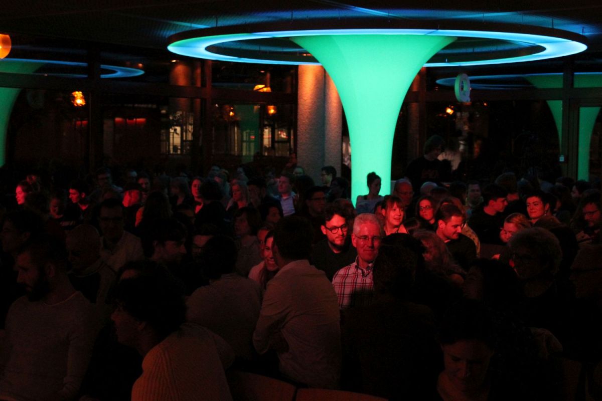 Ins Publikum fotografiert. Der Raum ist dunkel, aber ein großer Lichttrichter leuchtet grün-blau und auf einige Personen im Publikum scheint rotes Licht. Das Publikum diskutiert unterienander.